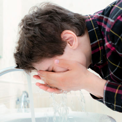 A boy splashing his face with water over a sink.